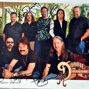 May include: A black and white photo of a band, The Doobie Brothers, with their signatures. The band members are standing in front of a wooden structure with a brown roof. The band members are wearing casual clothing. The band's name is written in a stylized font with a musical note.