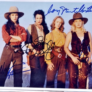 May include: A signed photograph of four women in western wear. The women are standing in a line, looking at the camera. The photo is signed by the women, with their names written in blue ink. The photo is set against a light blue sky background.