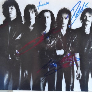May include: Black and white photograph of a rock band posing for a promotional shot. The five band members are wearing black leather jackets and pants. Each member has a signature written on the photo in blue and red ink.