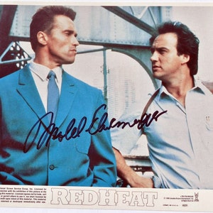 May include: A black and white photo of two men standing in front of a metal structure. The man on the left is wearing a blue suit and a blue tie. The man on the right is wearing a white shirt and a blue jacket. The photo is signed "Arnold Schwarzenegger". The title "RED HEAT" is printed in bold black letters at the bottom of the photo.