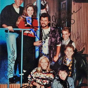 May include: A group of seven people, including a woman with short, grey hair, a woman with long, blonde hair, and five men, are standing in front of a wooden wall. They are all smiling and looking at the camera. The woman with short, grey hair is wearing a blue and black patterned dress. The woman with long, blonde hair is wearing a black and white patterned sweater. The men are wearing a variety of casual clothing. The wall behind them is decorated with wooden panelling and a blue and white striped pole.
