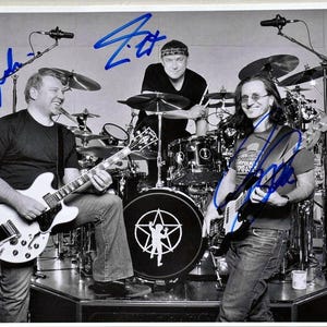 May include: A black and white photograph of the band Rush, featuring three musicians with guitars and a drum kit. The band members are smiling and posing. The image includes blue autographs.