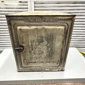 Puede incluir: Caja de pastel de metal vintage con puerta con bisagras. La caja es de color blanquecino con una ilustración descolorida en la parte delantera y el texto "SCHEFF'S CAKE BOX". La caja tiene un asa negra y adornos decorativos.