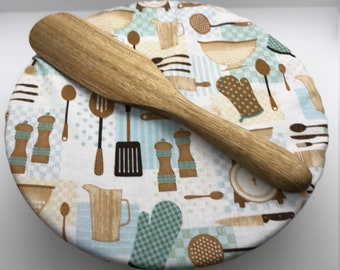 May include: A white fabric bowl cover with a blue and brown kitchen themed print. The cover has a wooden spatula on top.