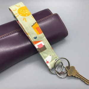 May include: A green and white fabric key chain with a sun, houses, and a white dog pattern. The key chain is attached to a silver key ring with a brass key.