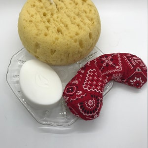Puede incluir: Una pastilla de jab&oacute;n blanca, una esponja de ba&ntilde;o amarilla y una m&aacute;scara para los ojos con estampado de bandana roja sobre un plato de vidrio transparente.