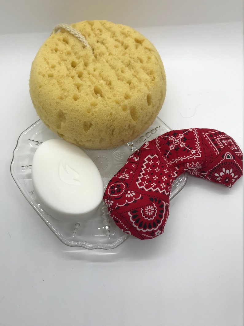 Puede incluir: Una pastilla de jab&oacute;n blanca, una esponja de ba&ntilde;o amarilla y una m&aacute;scara para los ojos con estampado de bandana roja sobre un plato de vidrio transparente.