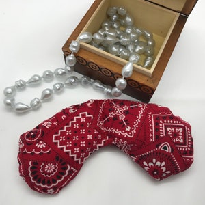 Puede incluir: Una almohada para los ojos de color rojo con un patr&oacute;n de bandana, con detalles en blanco y negro. La almohada tiene forma de media luna y est&aacute; sobre una superficie blanca. En el fondo, una caja de madera con un collar de perlas.