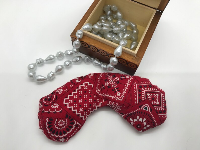 Puede incluir: Una almohada para los ojos de color rojo con un patr&oacute;n de bandana, con detalles en blanco y negro. La almohada tiene forma de media luna y est&aacute; sobre una superficie blanca. En el fondo, una caja de madera con un collar de perlas.