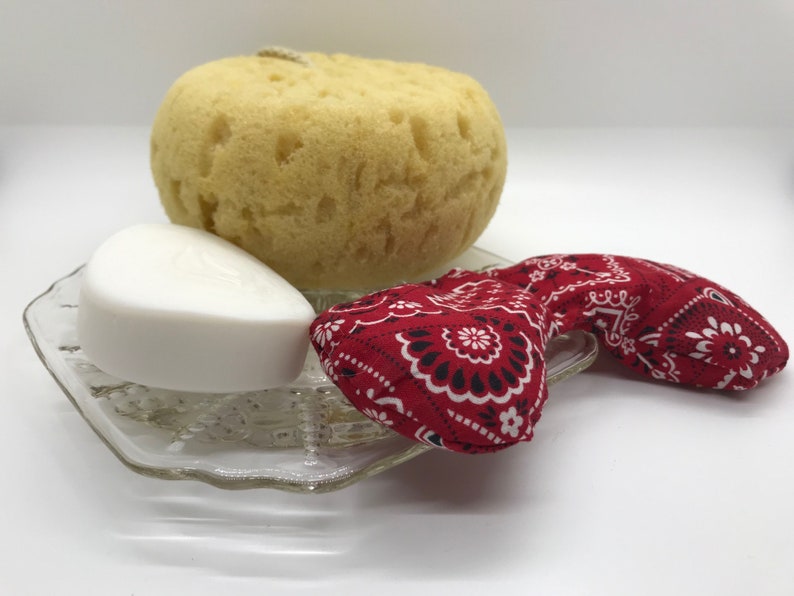 Puede incluir: Una pastilla de jab&oacute;n blanca, una esponja de ba&ntilde;o amarilla y una m&aacute;scara para los ojos de bandana roja en un plato de vidrio transparente.
