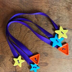 May include: Three purple ribbon bookmarks with colorful buttons. The buttons are shaped like stars and triangles. One star is yellow, one is blue, and one is orange. The triangles are orange.