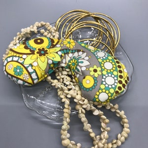 May include: An assortment of accessories, including a floral eye mask, gold bangles, and a seashell necklace, presented on a clear glass dish. The eye mask has yellow, green, and brown floral patterns. The bangles are thin and gold.