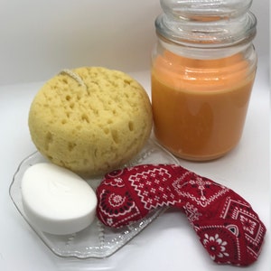 Puede incluir: Un set de regalo de spa con una esponja de ba&ntilde;o amarilla, una pastilla de jab&oacute;n blanca, una bandana roja y un tarro de vidrio de vela arom&aacute;tica naranja.