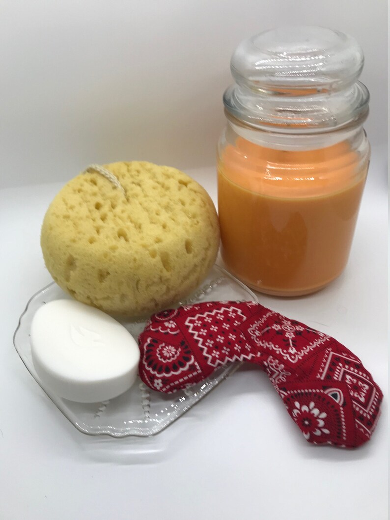 Puede incluir: Un set de regalo de spa con una esponja de ba&ntilde;o amarilla, una pastilla de jab&oacute;n blanca, una bandana roja y un tarro de vidrio de vela arom&aacute;tica naranja.