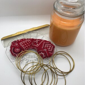 Puede incluir: Un conjunto de pulseras doradas en un plato de vidrio con una bandana roja. Un frasco de vidrio de cera de vela naranja est&aacute; en el fondo.