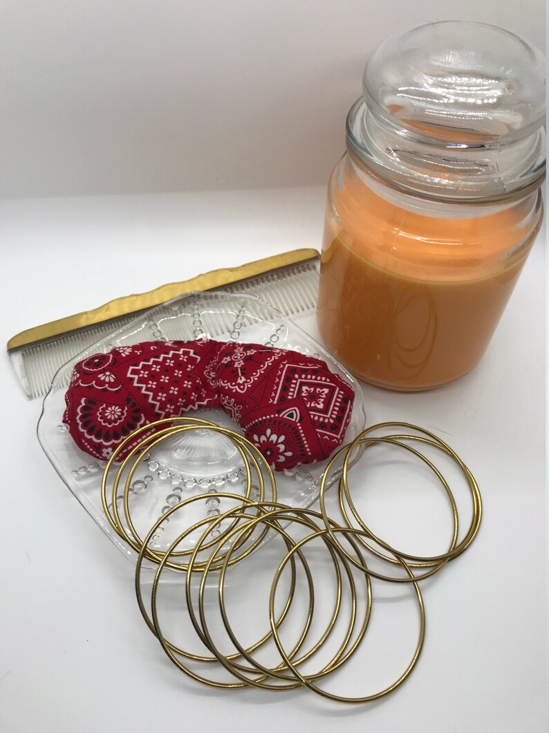 Puede incluir: Un conjunto de pulseras doradas en un plato de vidrio con una bandana roja. Un frasco de vidrio de cera de vela naranja est&aacute; en el fondo.