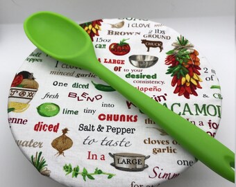 May include: A white fabric pot lid with a green silicone spoon on top. The lid is printed with a recipe for guacamole, including ingredients like brown ground chuck, diced tomatoes, lime, and garlic. The text also includes measurements in ounces and pounds.