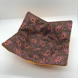 May include: A square-shaped fabric bowl with a paisley pattern in shades of brown, red, and yellow. The bowl is lined with a solid yellow fabric.
