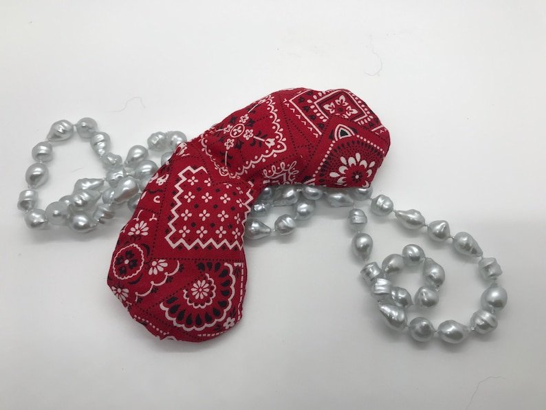 Puede incluir: Una m&aacute;scara para los ojos de tela de bandana roja con un patr&oacute;n paisley blanco y negro, descansando sobre una hebra de perlas gris plateado.