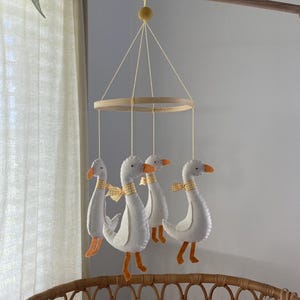 Könnte beinhalten: Ein Baby-Mobile mit vier weißen Gänse-Plüschtieren mit orangefarbenen Schnäbeln und Füßen sowie gelb karierten Schleifen. Die Gänse hängen an einem Holzring und sind an gelben Schnüren aufgehängt. Oben befinden sich eine Holzstange und ein gelber Pompon.