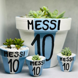 May include: Four white ceramic succulent pots with blue accents. The largest pot has the words "Messi 10" painted on it. The three smaller pots have the words "Messi 10" painted on them in a similar style.