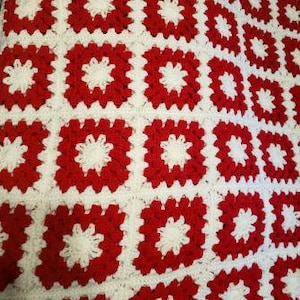 Könnte beinhalten: Eine rot-weiße Häkeldecke mit quadratischem Muster. Die Decke ist aus Garn gefertigt und hat eine weiche, strukturierte Oberfläche.