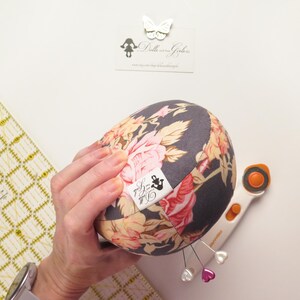 May include: A floral print pincushion with pink and peach roses on a dark gray background. The pincushion has a small label with text and is held near a rotary cutter, pins, and a quilting ruler.