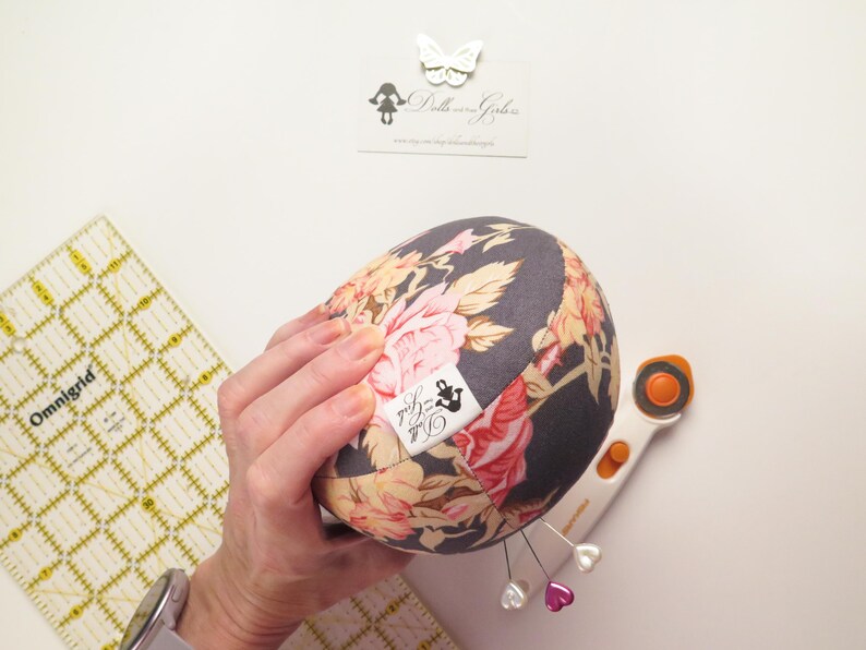 May include: A floral print pincushion with pink and peach roses on a dark gray background. The pincushion has a small label with text and is held near a rotary cutter, pins, and a quilting ruler.