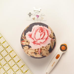 May include: A round pincushion with a large pink rose design, surrounded by tan and green leaves, sits next to a yellow grid ruler and a white rotary cutter. The pincushion has pins with heart and butterfly toppers.