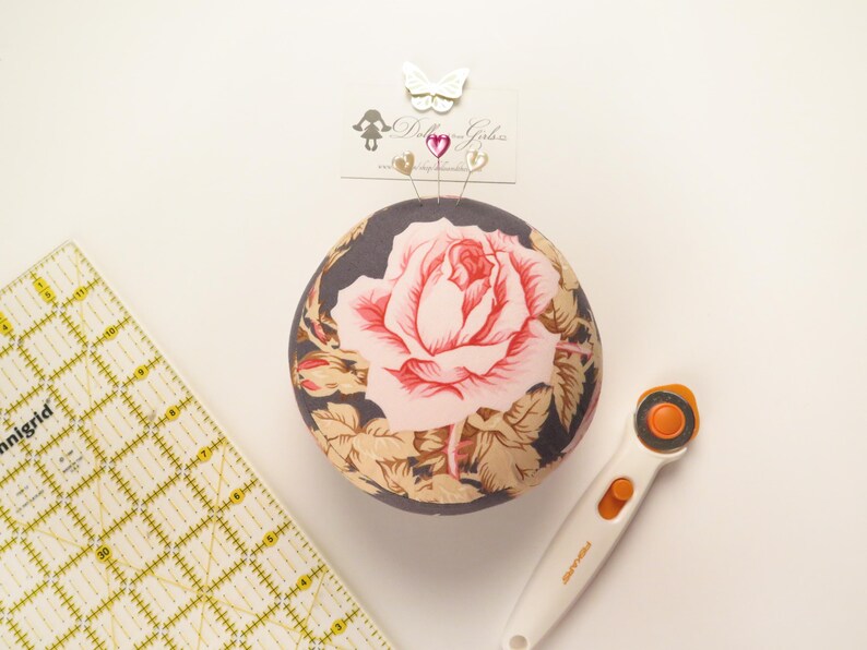 May include: A round pincushion with a large pink rose design, surrounded by tan and green leaves, sits next to a yellow grid ruler and a white rotary cutter. The pincushion has pins with heart and butterfly toppers.