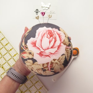 May include: A round pincushion with a large pink rose design on a dark gray background. The pincushion has decorative pins and a butterfly embellishment. A rotary cutter and Omnigrid ruler are also visible.