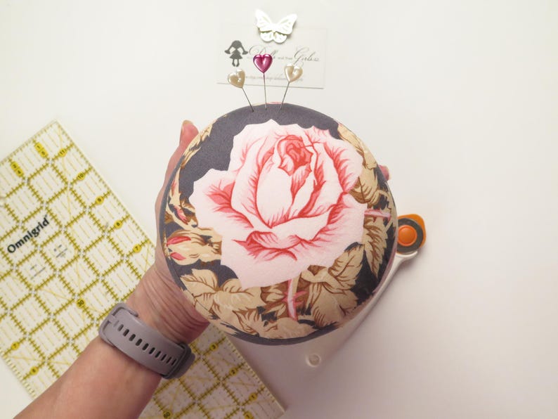 May include: A round pincushion with a large pink rose design on a dark gray background. The pincushion has decorative pins and a butterfly embellishment. A rotary cutter and Omnigrid ruler are also visible.