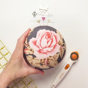 May include: A round pincushion with a large pink rose design on a dark background. Three decorative pins are inserted into the top. A rotary cutter and a ruler are also visible.