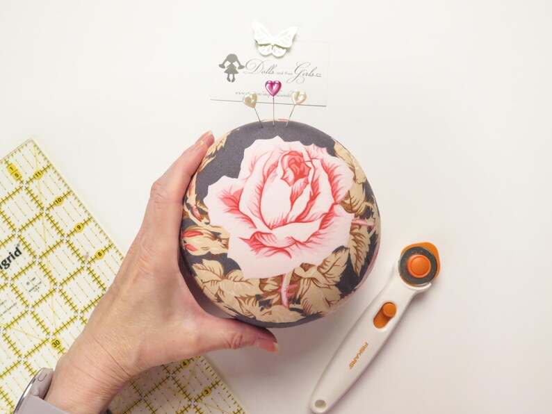 May include: A round pincushion with a large pink rose design on a dark background. Three decorative pins are inserted into the top. A rotary cutter and a ruler are also visible.
