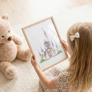 May include: A framed watercolor print of a castle with tall blue-roofed towers and a central clock face, displayed in a light wood frame. The artwork features soft pastel tones and green foliage at the base, presented as a decorative wall art piece.