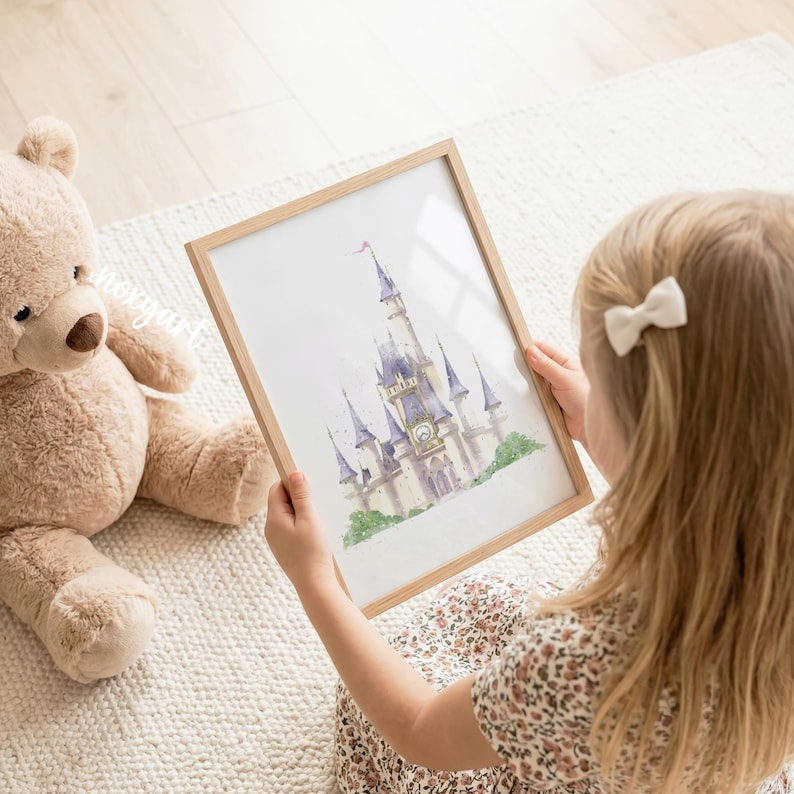 May include: A framed watercolor print of a castle with tall blue-roofed towers and a central clock face, displayed in a light wood frame. The artwork features soft pastel tones and green foliage at the base, presented as a decorative wall art piece.