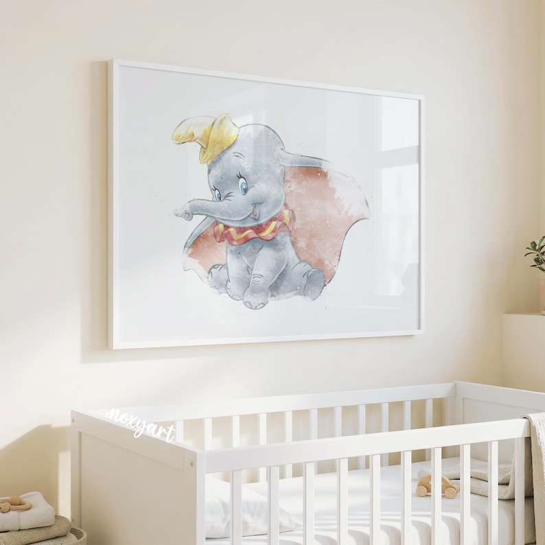 May include: A framed watercolor print of Dumbo the elephant, featuring a gray elephant with a yellow hat, red and yellow collar, and large pink ears. The artwork is displayed in a white frame above a white crib, creating a nursery theme.