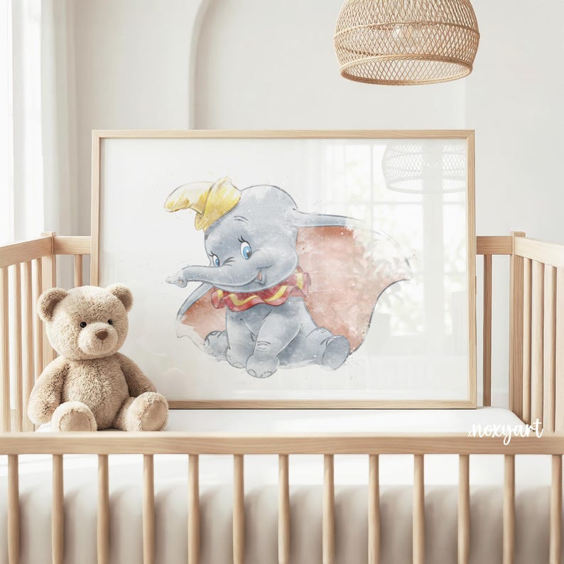 May include: A framed watercolor print of Dumbo the elephant, featuring a gray elephant with a yellow hat and a red and pink cape. The artwork is displayed in a light wood frame, suitable for a nursery or children's room. A teddy bear sits in a wooden crib.