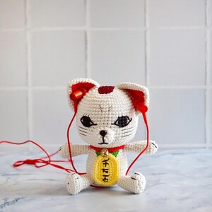 May include: A handmade white crochet cat figurine with red accents, wearing red headphones. The cat has black eyes and a yellow tag with Japanese characters. The cat is sitting with its legs extended.
