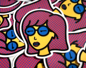 Könnte beinhalten: Ein Stapel bunter Aufkleber mit einer Comic-Frau mit roten Haaren und blauen Brillen. Die Aufkleber haben einen Pop-Art-Stil mit einem roten und blauen Punktmuster im Hintergrund.