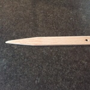 May include: A wooden tool with a pointed tip and a hole near the rounded end. The tool is approximately 6 inches long.