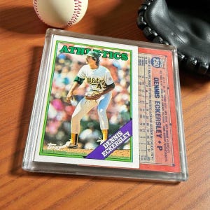 May include: A baseball card of Dennis Eckersley in a protective case. The card features Eckersley in an Athletics uniform. A baseball and a black baseball glove are in the background. The card is from the Topps brand.