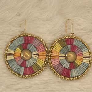 May include: A pair of colorful, round earrings. Each earring features a circular design with a gold-colored center and a beaded gold-colored border. The interior design is a radial pattern of pastel colors, including pink, blue, yellow, and green.