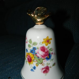 Könnte beinhalten: Eine weiße Porzellan-Glocke mit einem goldenen Vogelknauf und einem Blumenmuster. Die Glocke ist mit rosa, gelben, blauen und roten Blumen verziert.