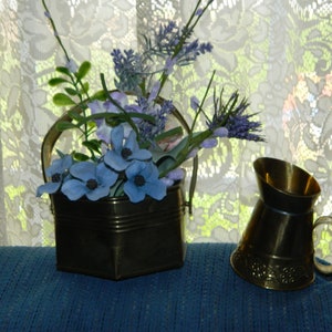 Puede incluir: Un cubo de latón con asa, lleno de flores artificiales azules y moradas. El cubo está sobre una superficie azul, junto a una jarra de latón con asa.