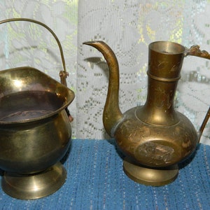 Puede incluir: Dos recipientes antiguos de latón. Uno es un pequeño cubo con asa, y el otro es una jarra decorativa con un pico largo y diseños florales grabados. Ambos están sobre una superficie azul.