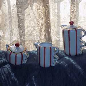 May include: A light blue ceramic tea set with red vertical stripes and gold trim. The set includes a teapot, creamer, and sugar bowl, all with a whimsical, wavy handle design. The teapot has a red knob on the lid.