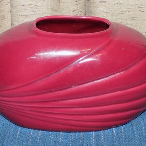 Puede incluir: Un jarrón de cerámica rojo oscuro con un acabado liso y brillante. El jarrón tiene una forma ovalada redondeada con una abertura ancha en la parte superior. La superficie del jarrón presenta una serie de líneas curvas en relieve que crean un diseño fluido y orgánico.