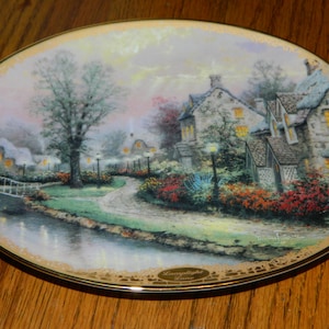 May include: A decorative ceramic plate featuring a painting of a quaint village scene at twilight. The plate has a gold rim and a gold inscription that reads "Moonlight Lace".