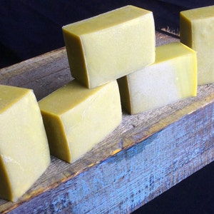 May include: Five bars of handmade soap, all a pale yellow-green color, are arranged on a rustic wooden surface. The surface is painted a faded blue color.